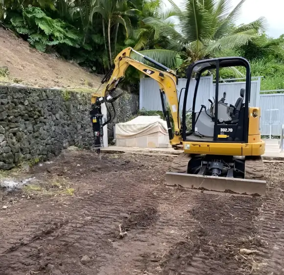 kona-heavy-equipment-landscaping A yellow compact excavator with a jackhammer attachment digs into the dirt beside a lava rock wall, illustrating the need for heavy equipment for big island landscape projects.