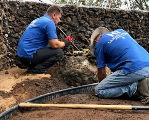 Kailua-Kona landscape workers in blue shirts install edging in dark soil next to a volcanic rock wall, highlighting Hawaii lava vs soil challenges.