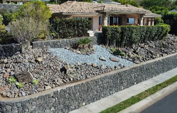 kona-dry-climate Drought-resistant garden setup showing tiered volcanic rock walls and smooth blue stones outside a house, typical of Kona landscaping.