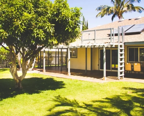 Yellow home with a covered lanai and solar panels sits behind a broad shade tree and palm, an example of an energy-saving landscape design in Kailua-Kona.